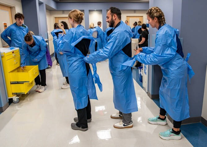 Students in a simulated hospital help each other put on medical gowns